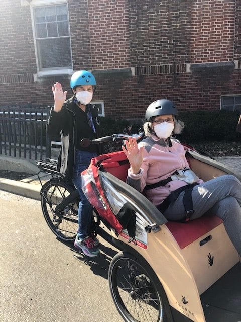 Two individuals wearing helmets and masks enjoy riding a tricycle outdoors.