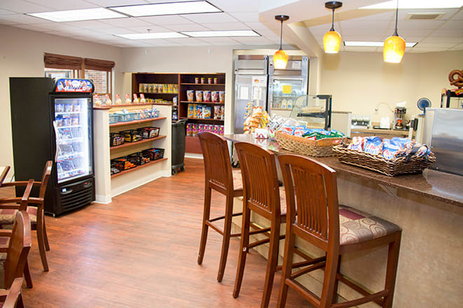 A cozy cafe interior featuring a seating area with high wooden chairs, a counter with baked goods, and shelves stocked with various items.
