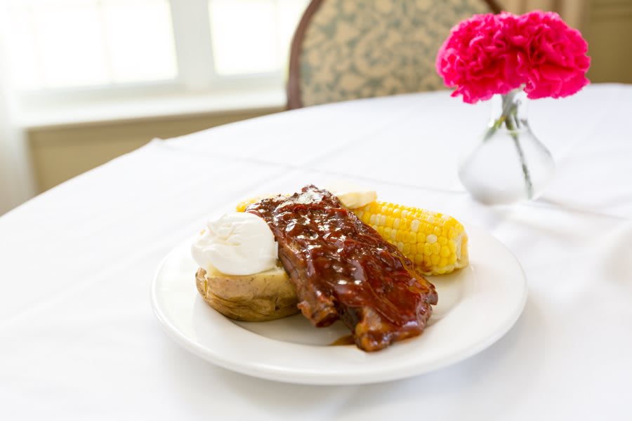 A delicious meal consisting of BBQ ribs, baked potato with sour cream, and corn on the cob served on a white plate.