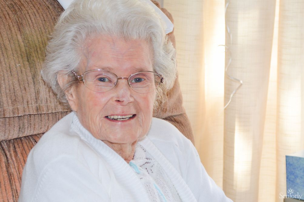 An elderly woman with white hair and glasses smiling warmly indoors.
