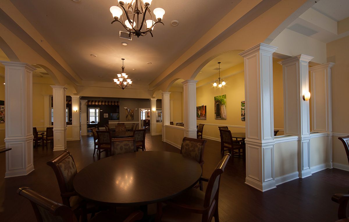 A spacious dining room illuminated by elegant chandeliers, featuring a large table surrounded by chairs and decorative columns.