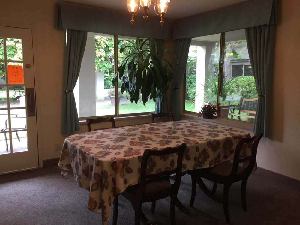 A cozy dining room with a table covered by a patterned cloth, surrounded by chairs and illuminated by a chandelier. Large windows allow daylight to brighten the space.