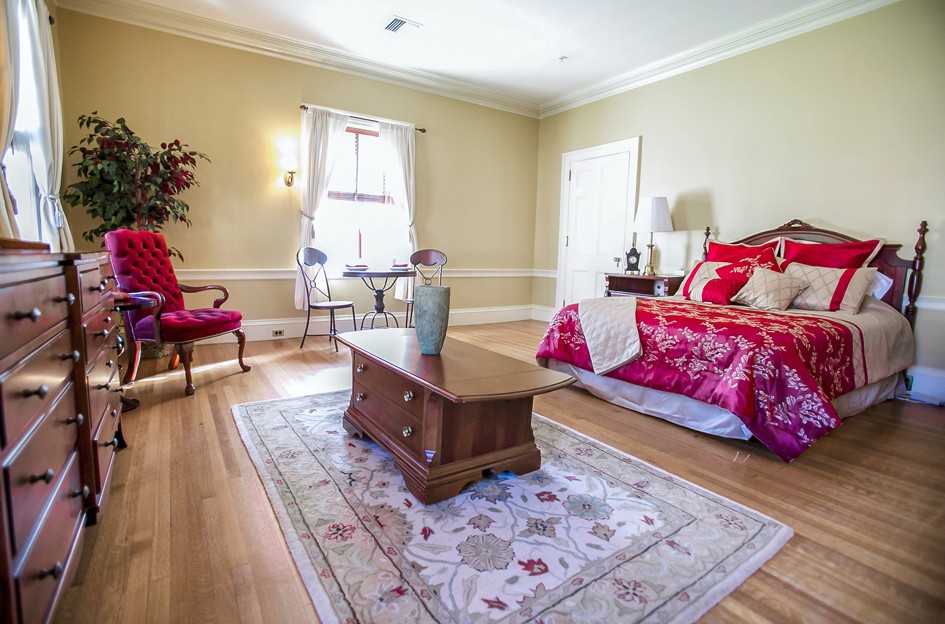 A cozy bedroom with a wooden floor, featuring a bed adorned with red bedding, a central rug, and elegant furnishings.