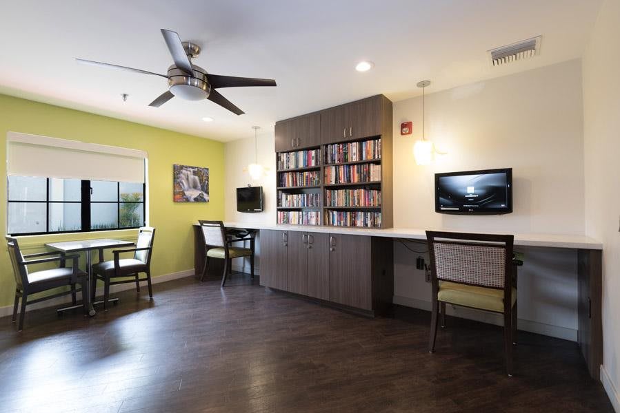 A cozy reading and study area featuring a bookshelf, two tables with chairs, a ceiling fan, and a television on the wall.