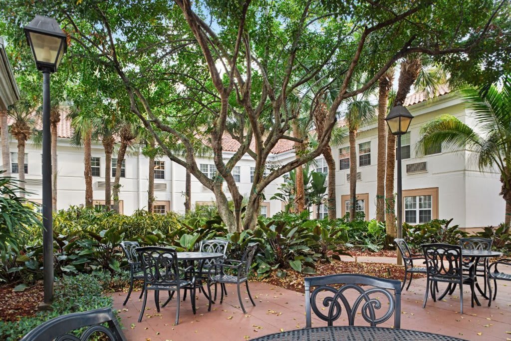 A serene outdoor patio area with tables and chairs surrounded by lush greenery and trees, adjacent to a building.