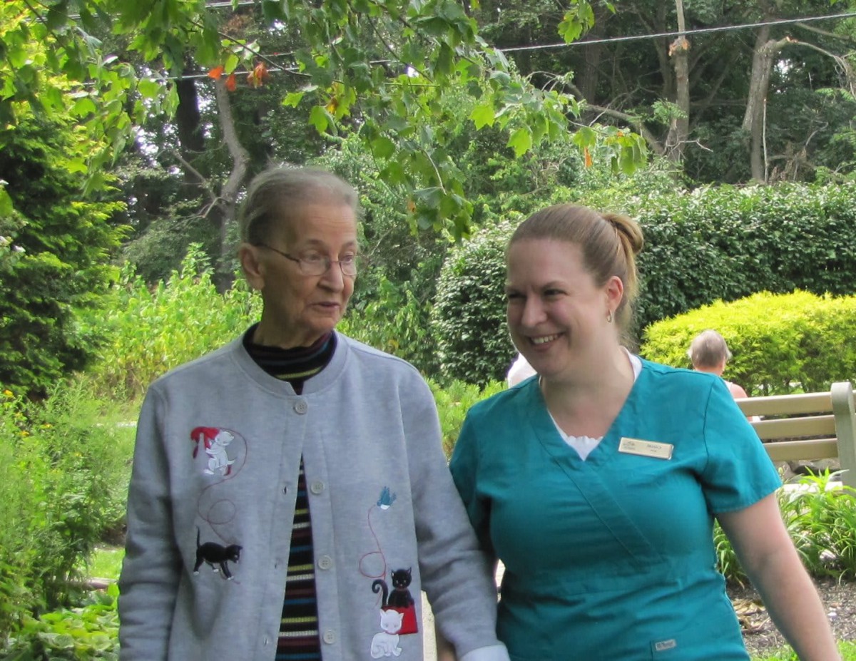 An elderly person walking with a caregiver in a park setting.