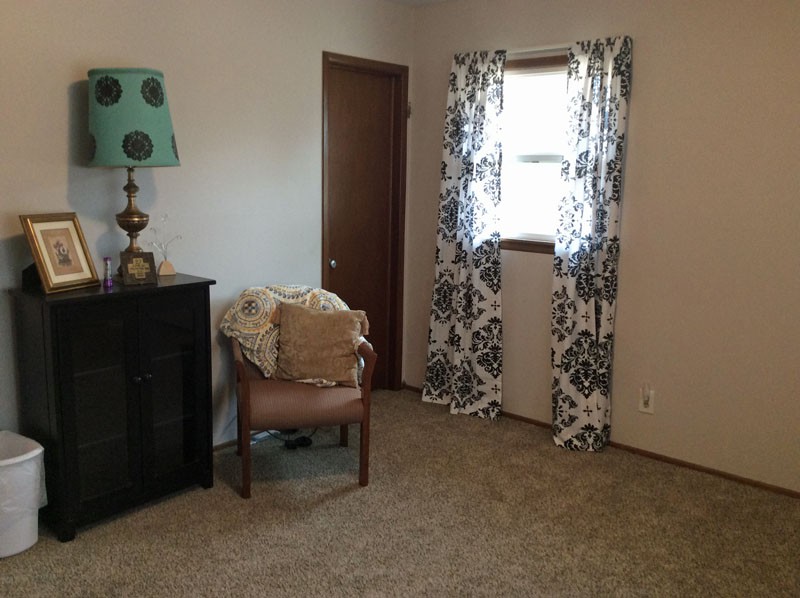 A cozy room featuring a chair with a decorative pillow, a lamp on a small table, and patterned curtains by the window.