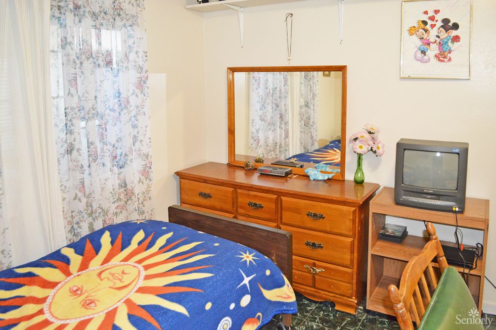 A cozy bedroom with a sun-themed comforter, wooden dresser with a mirror, and a small TV on a stand.