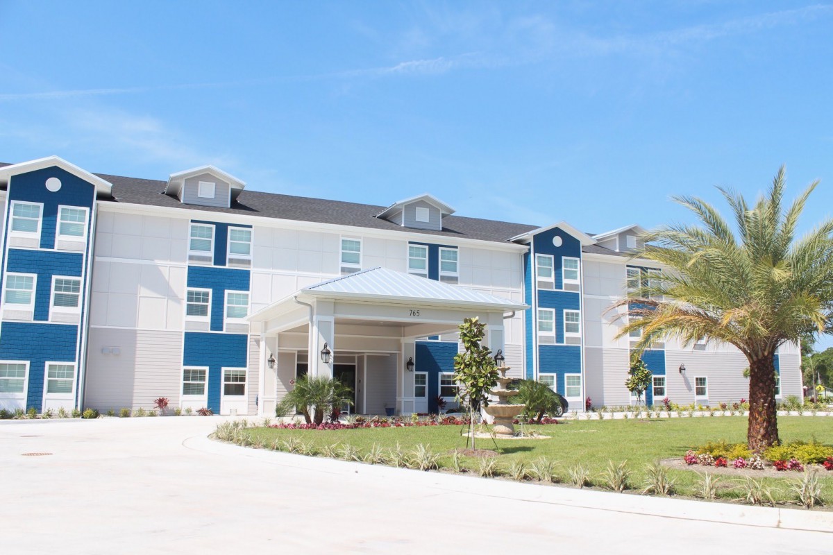 A modern hotel building with a welcoming entrance, surrounded by landscaped greenery and set against a clear blue sky.