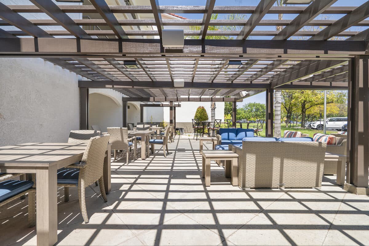 A modern outdoor patio with tables and chairs under a pergola, bathed in sunlight.