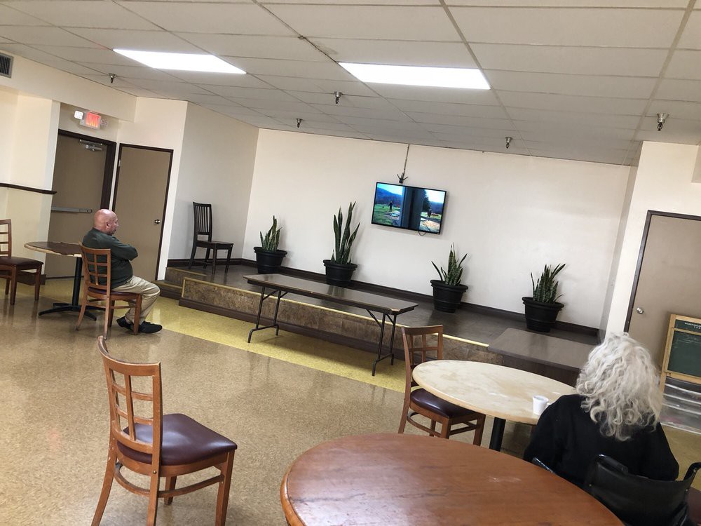 An empty waiting area with tables, chairs, a small stage with plants, and a television on the wall.