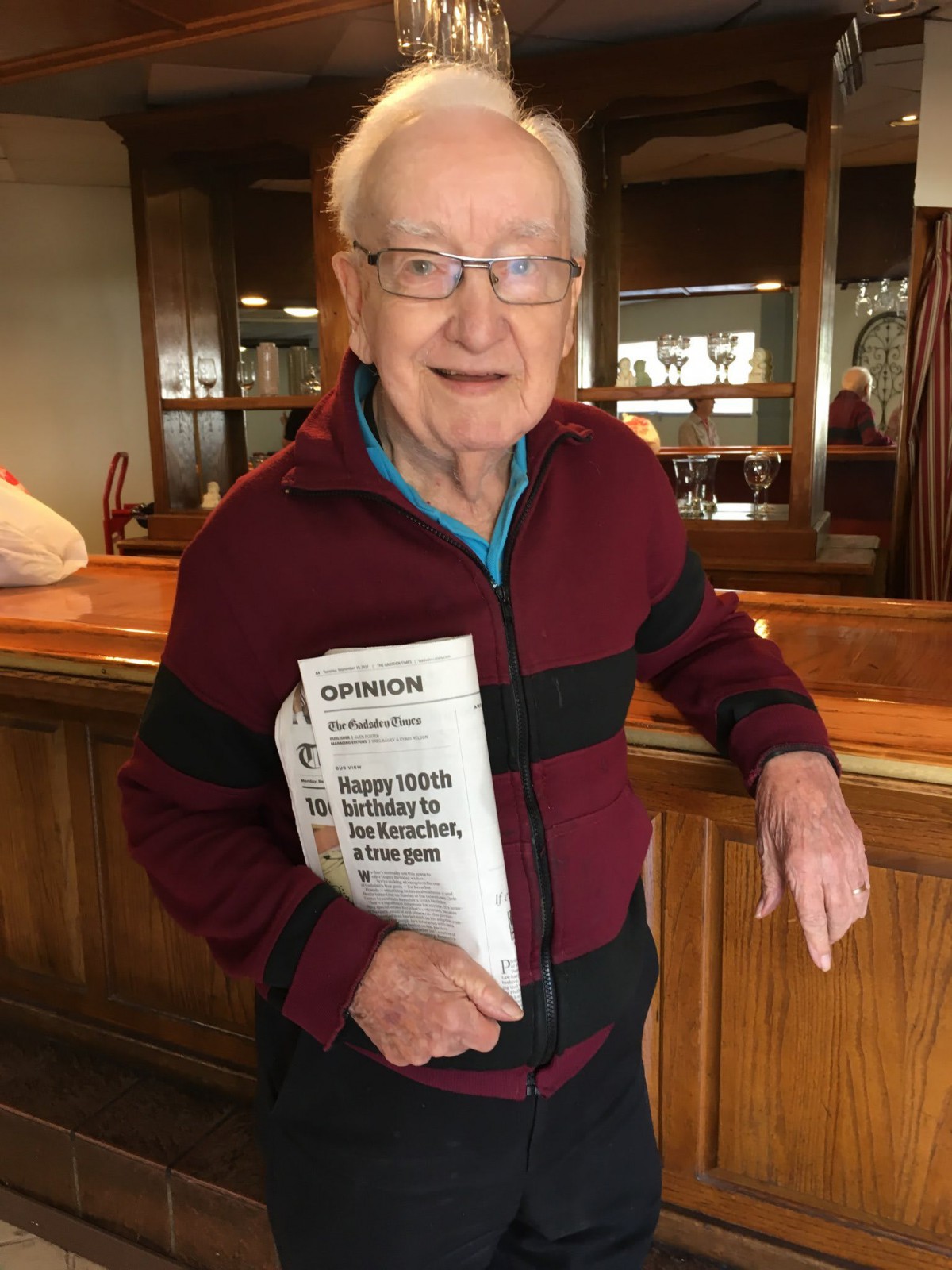 An elderly man smiling indoors while holding a newspaper.