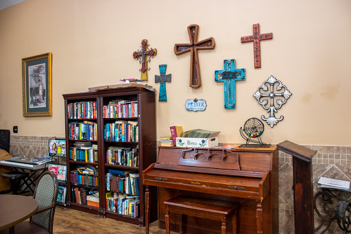A cozy corner of a room featuring a wooden bookshelf filled with books, a piano, and an array of decorative crosses on the wall.