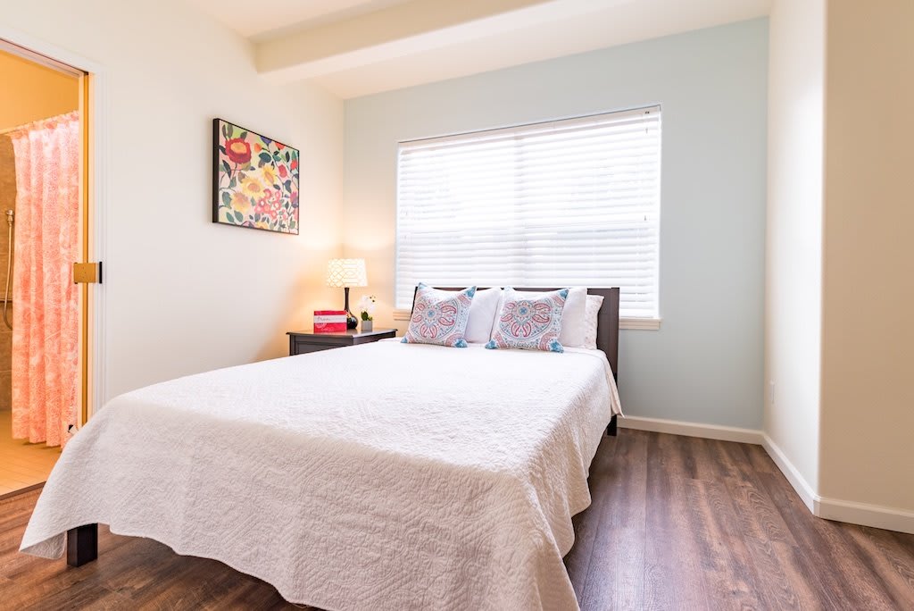 A bright and cozy bedroom with a neatly made bed, bedside table, and artwork on the wall.