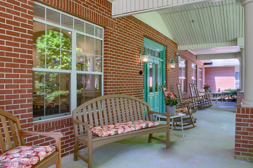 A cozy porch with wooden benches, rocking chairs, and decorative pillows against a brick wall.