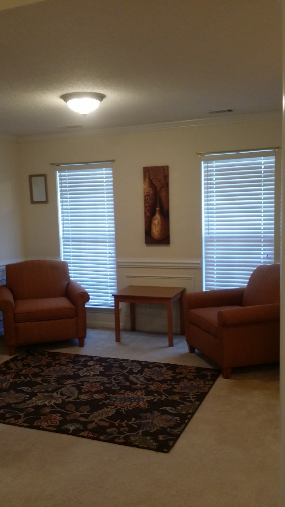 A cozy living room with two armchairs, a small table, and two windows with blinds.
