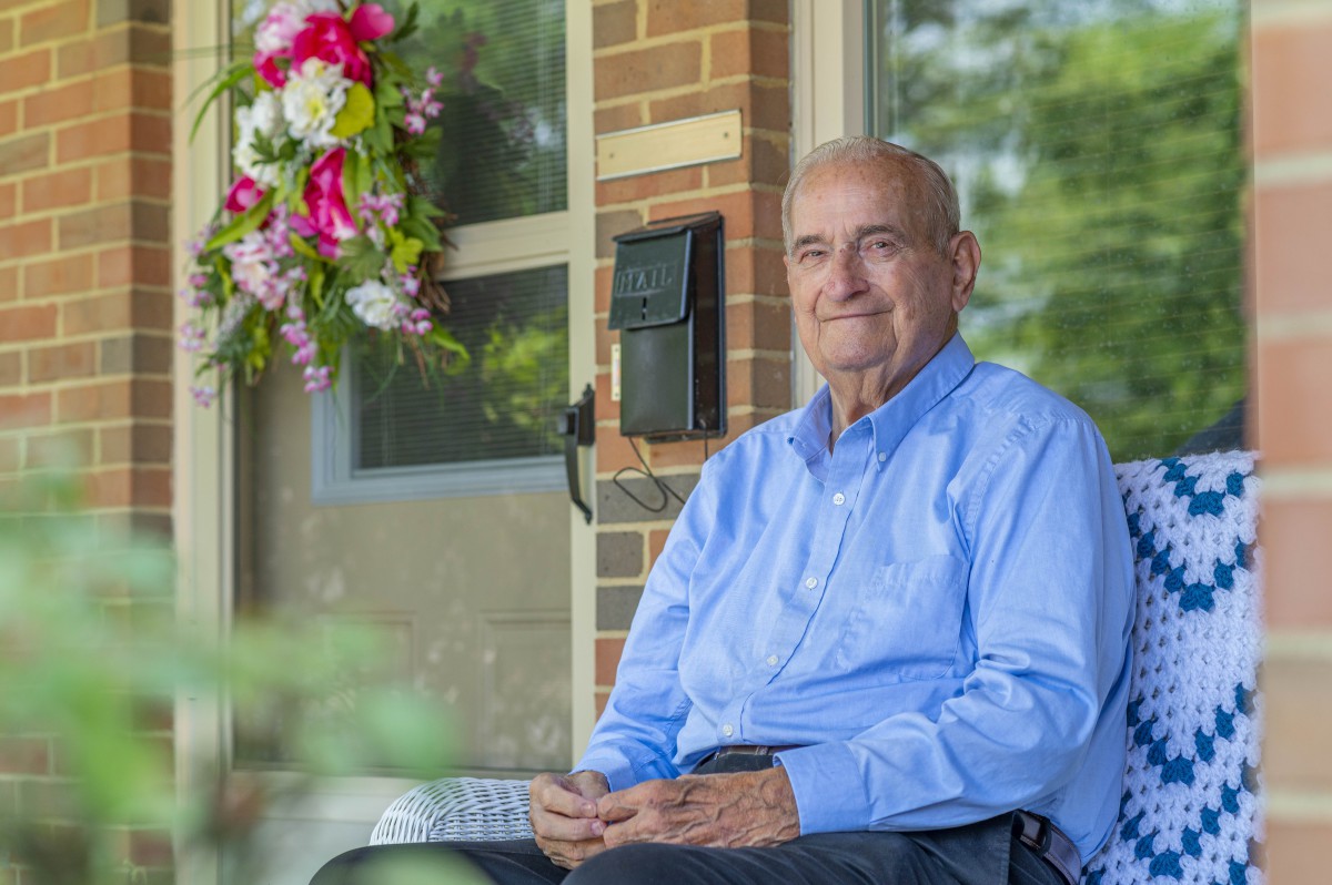 An elderly man sitting on a porch with a flower arrangement and a brick wall behind him.