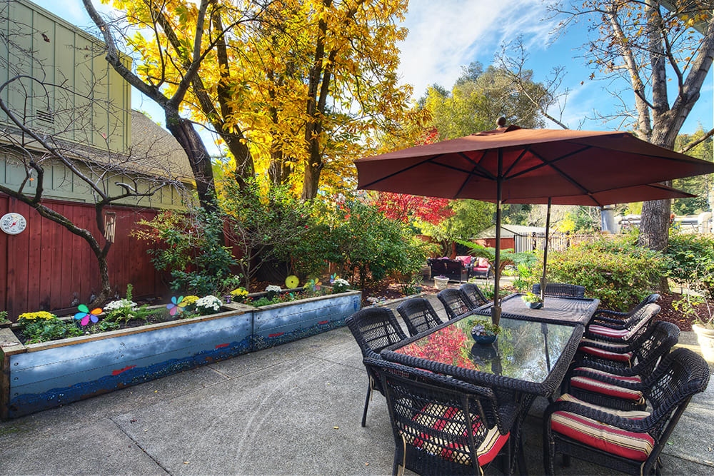 A charming outdoor patio with tables and chairs, surrounded by lush garden greenery and autumn trees.
