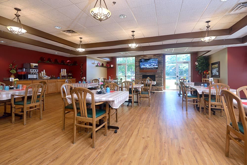 A cozy dining area with wooden floors, tables, and chairs, lit by chandeliers and a large window view.