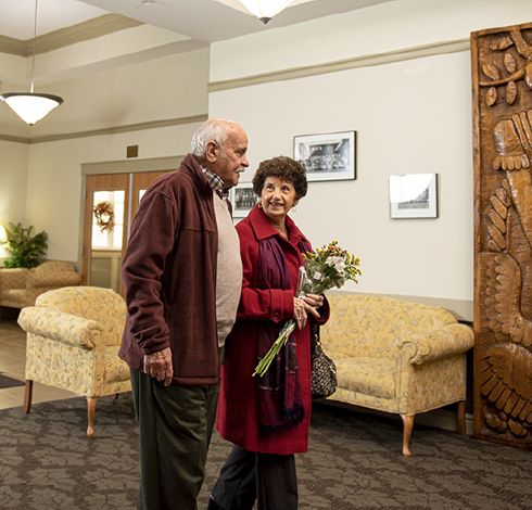An elderly couple walks together indoors, with the woman holding a bouquet of flowers.