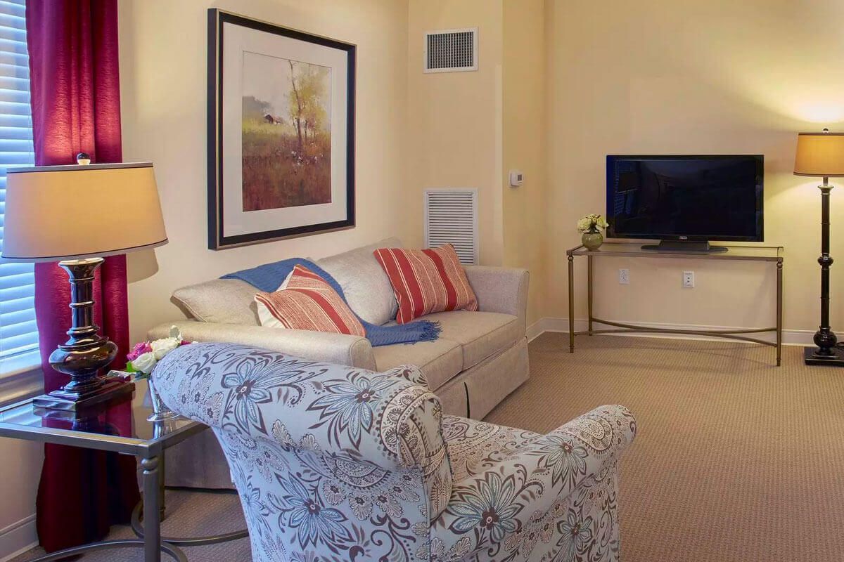 A cozy living room with a sofa, armchair, and television, featuring artwork on the wall.