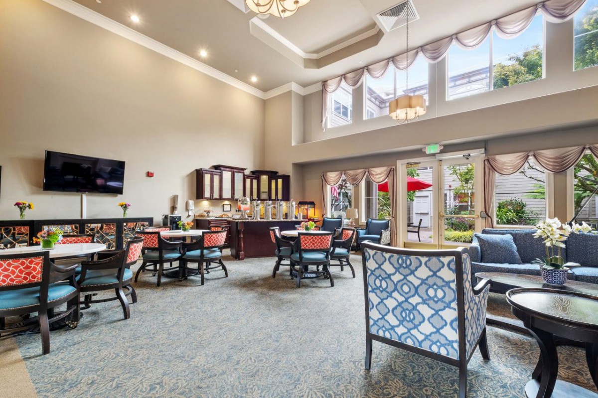 A spacious and well-lit dining area with elegant decor, featuring multiple tables and chairs, large windows, and a chandelier.