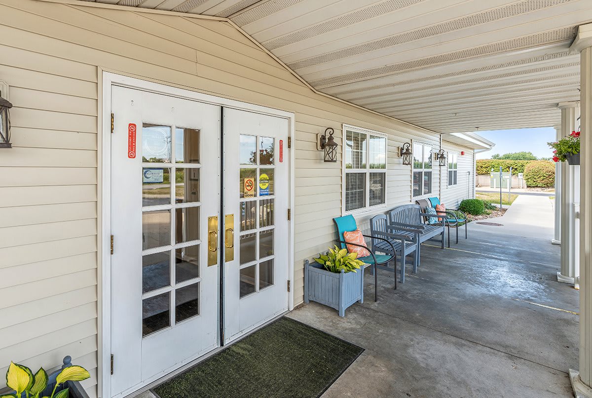 A welcoming building entrance with large glass doors, benches, and flower decorations under an awning.