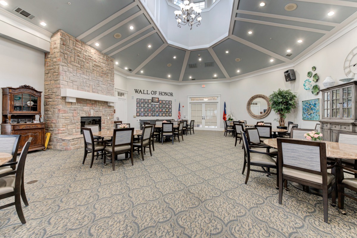 A spacious dining area with multiple tables and chairs, featuring a chandelier, fireplace, and a 'Wall of Honor' display.