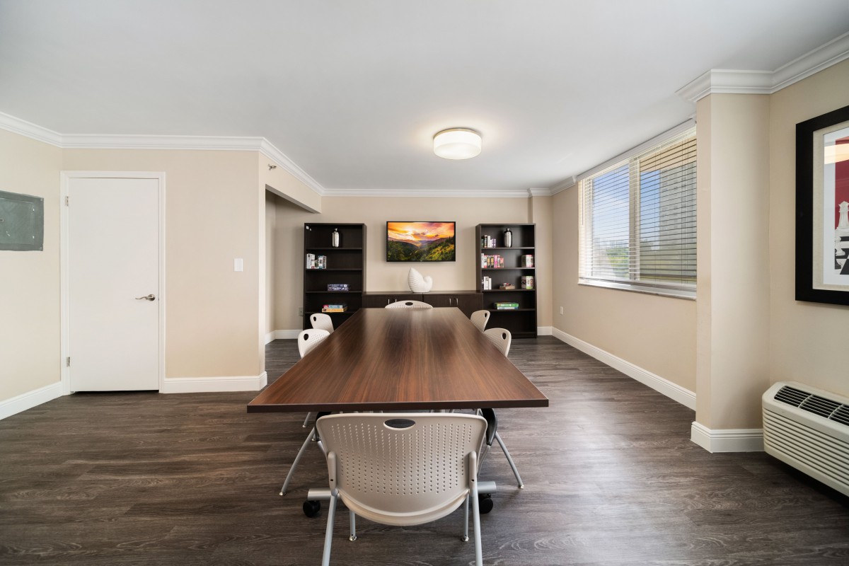 A modern meeting room with a large table and chairs, featuring bookshelves and a window.