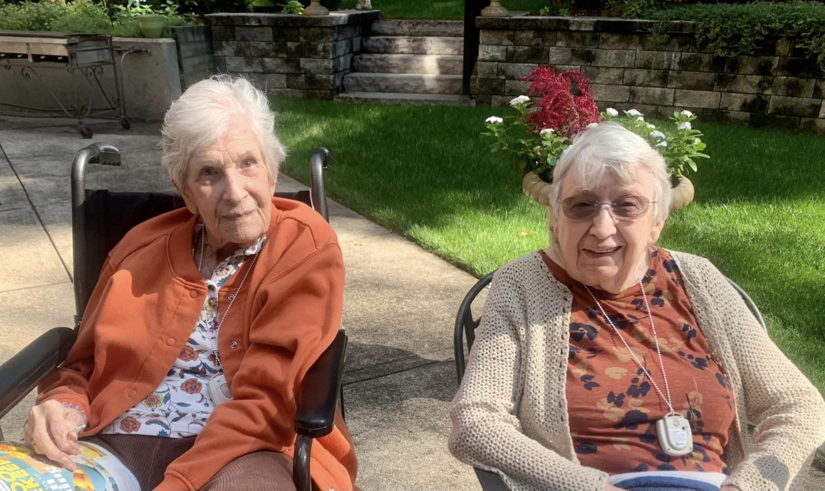 Two elderly women sitting outdoors in wheelchairs on a sunny day.