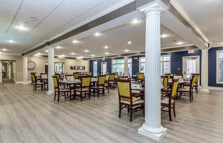 A spacious dining room with multiple tables and chairs, featuring elegant columns and bright lighting.