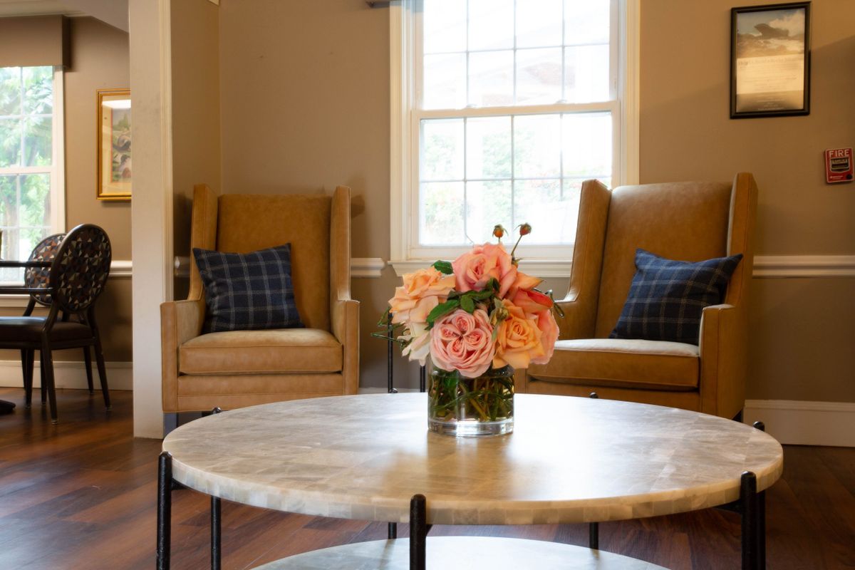 A cozy living room featuring two armchairs, a round coffee table with a bouquet of flowers, and a window letting in natural light.