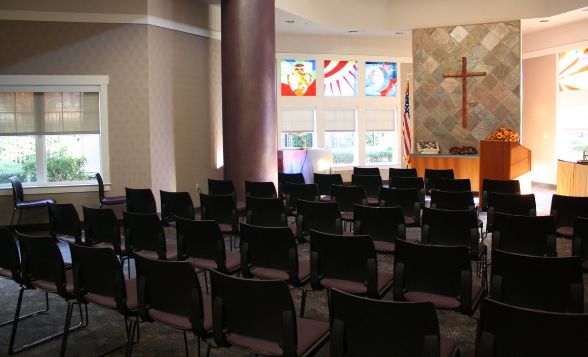 An empty chapel with rows of chairs, a cross on the wall, and stained glass windows.