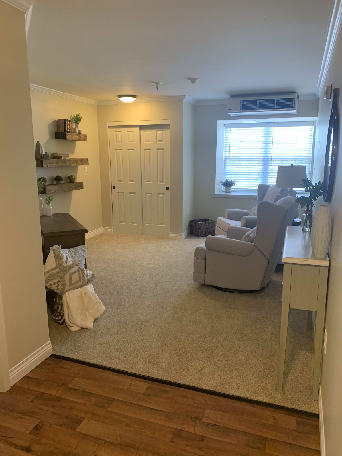 A cozy living room with carpeted flooring, featuring comfortable seating and natural light streaming through a large window.