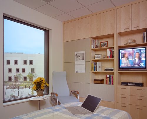 A cozy hospital room with a view, featuring a bed, chair, bookshelf, and television.