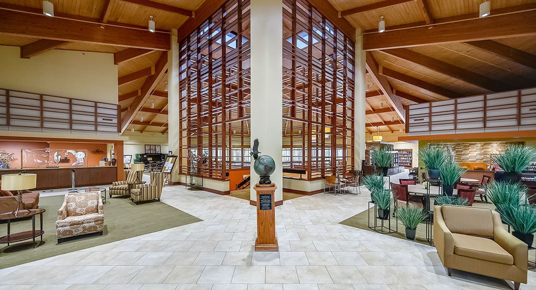 Modern library interior with wooden beams and large glass windows, featuring a central statue.