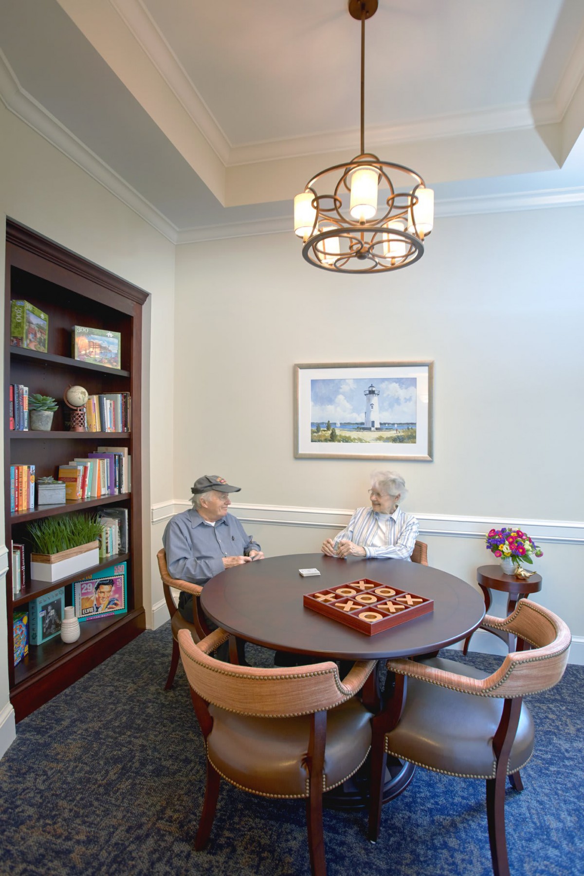 Elderly individuals sitting in a cozy room with a chess board on the table and a bookshelf nearby.