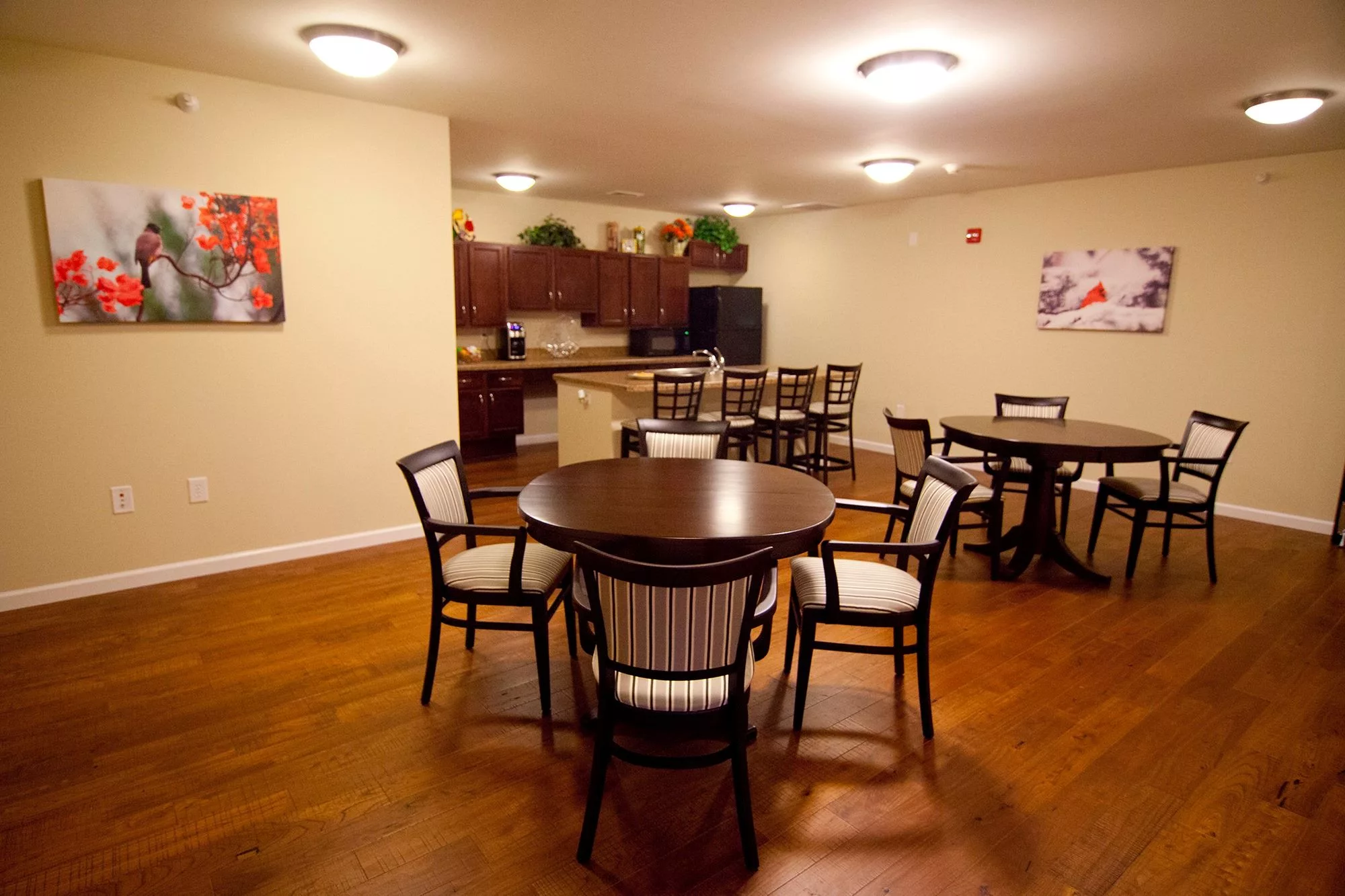 A cozy dining area with wooden flooring, featuring multiple tables and chairs adjacent to a kitchen.
