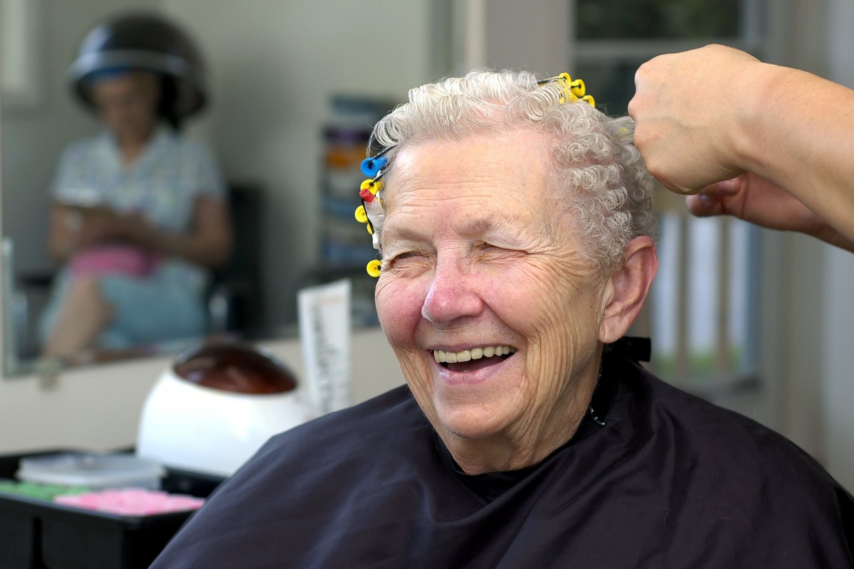 An elderly woman smiles while having her hair done at a salon.