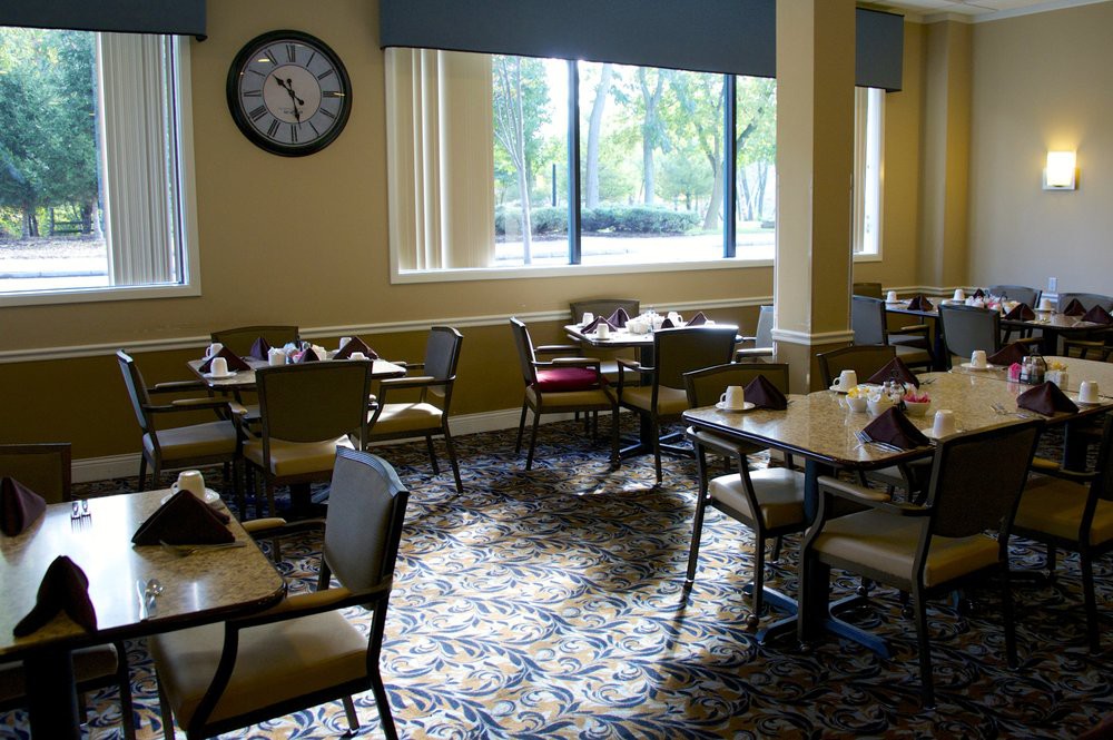 An empty dining room with tables and chairs, featuring a large clock on the wall and natural light from windows.