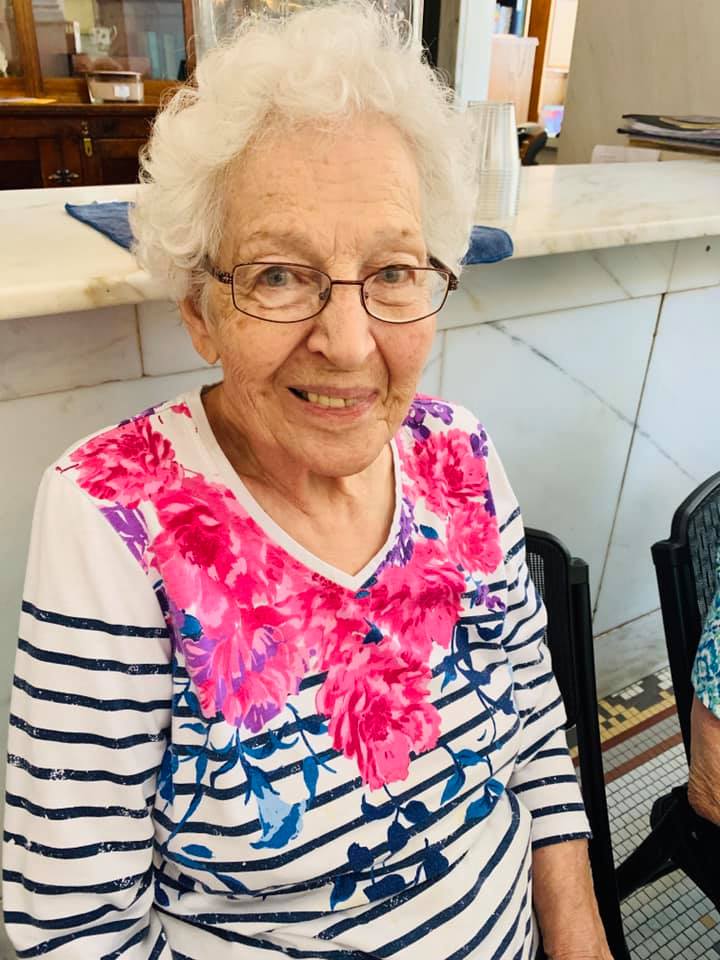 An elderly woman with glasses wearing a striped shirt with floral design, smiling indoors.