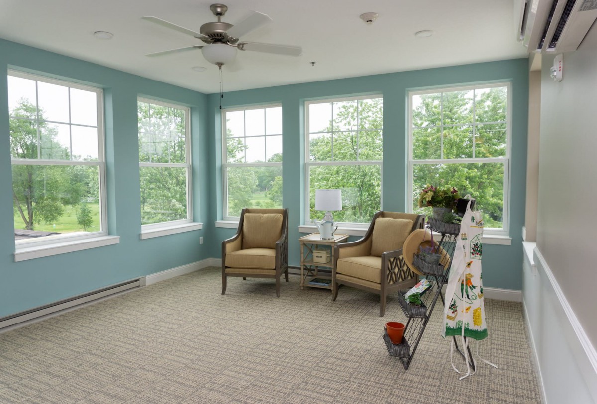 A bright sunroom with large windows, featuring two chairs, a small table, and an easel.