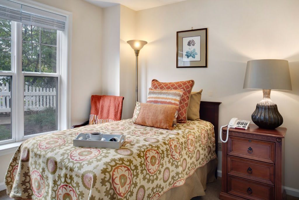 A cozy bedroom with a single bed adorned with patterned bedding and decorative pillows, featuring a lamp, a nightstand, and a window providing natural light.