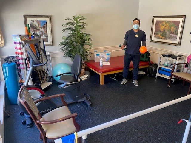 A healthcare worker wearing a mask in a physical therapy room with various exercise equipment.