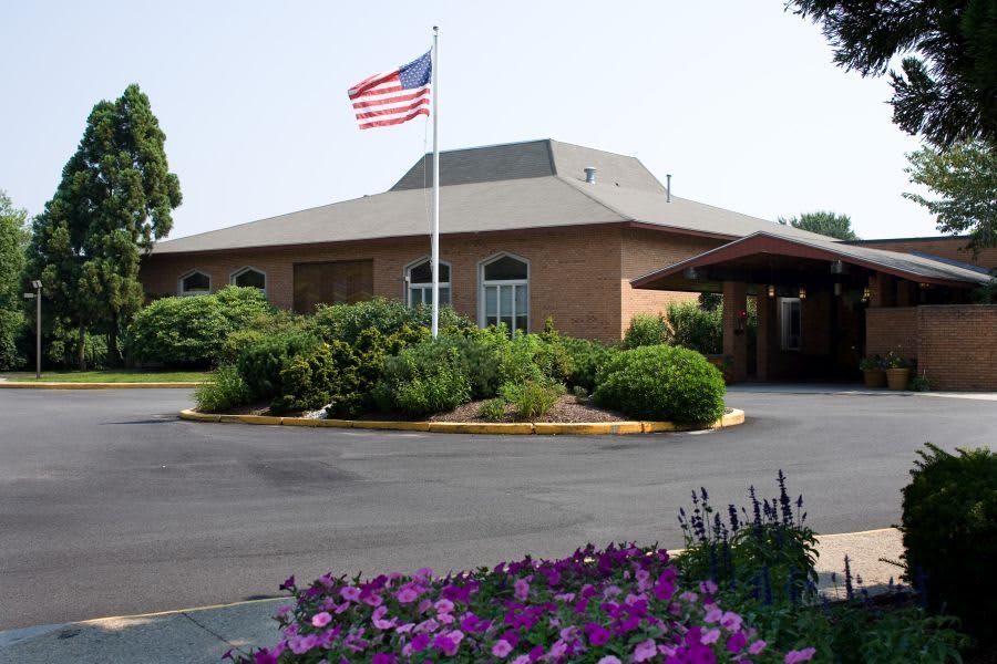 A brick building with an American flag flying in front, surrounded by lush landscaping and colorful flowers.
