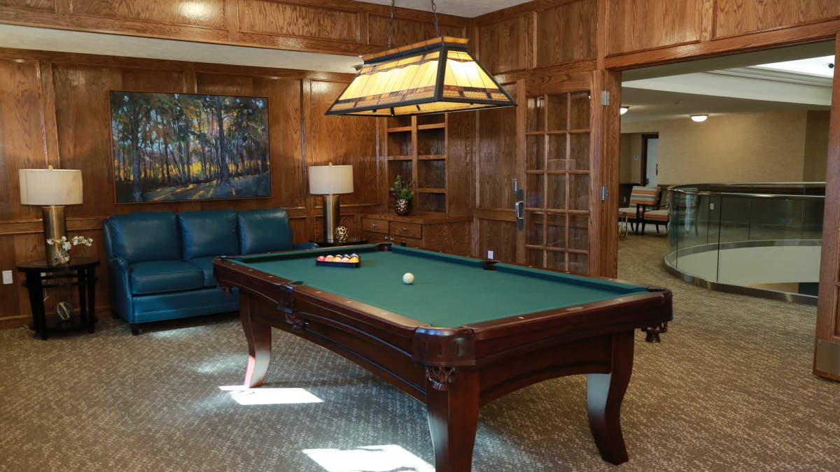 A cozy game room featuring a billiards table with a hanging lamp above it, surrounded by wood-paneled walls and comfortable seating.
