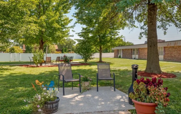 A peaceful backyard patio area with two chairs, surrounded by a well-maintained lawn and lush trees.
