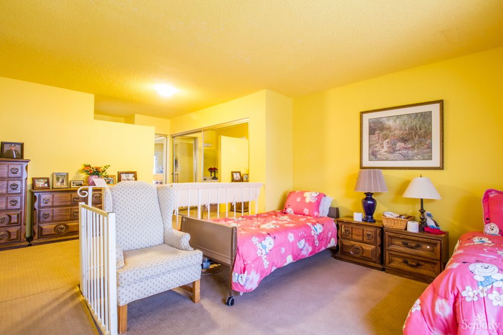 A cozy bedroom with yellow walls, featuring a single bed covered in a pink blanket, an armchair, and a nightstand with a lamp.