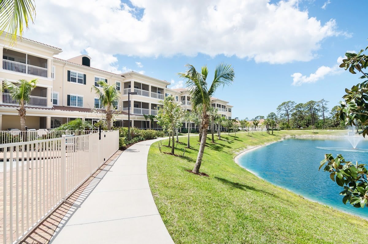 A scenic view of an apartment complex by a lake, surrounded by lush greenery and palm trees under a cloudy sky.