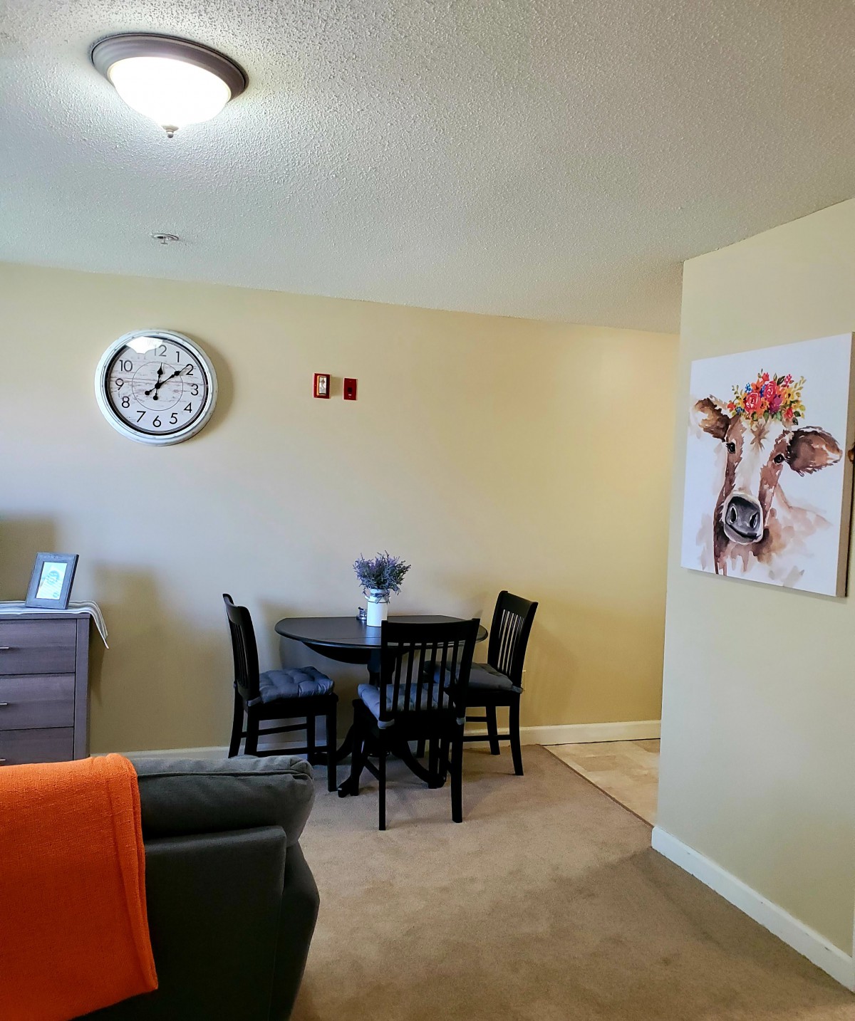 A cozy dining area with a round table and chairs, adorned by a cow painting and wall clock.